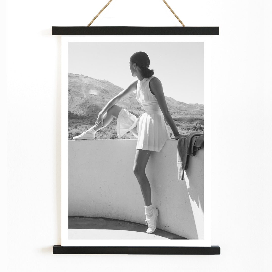 Fashion model in tennis outfit seated on wall looking at mountains by Toni Frissell poster artwork, with wooden magnetic frame on white background
