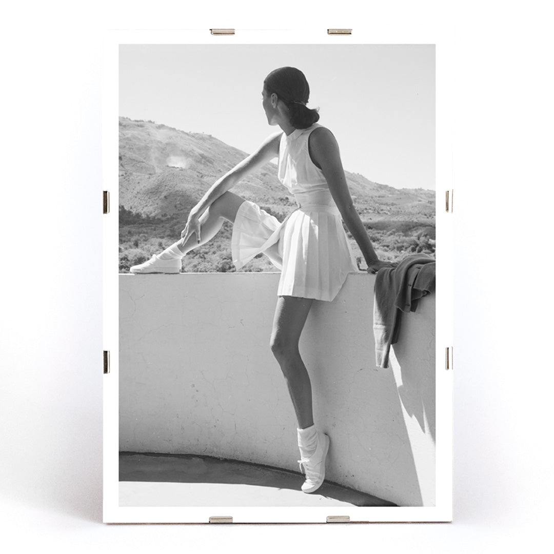 Fashion model in tennis outfit seated on wall looking at mountains by Toni Frissell poster artwork, framed and displayed in modern living room decor