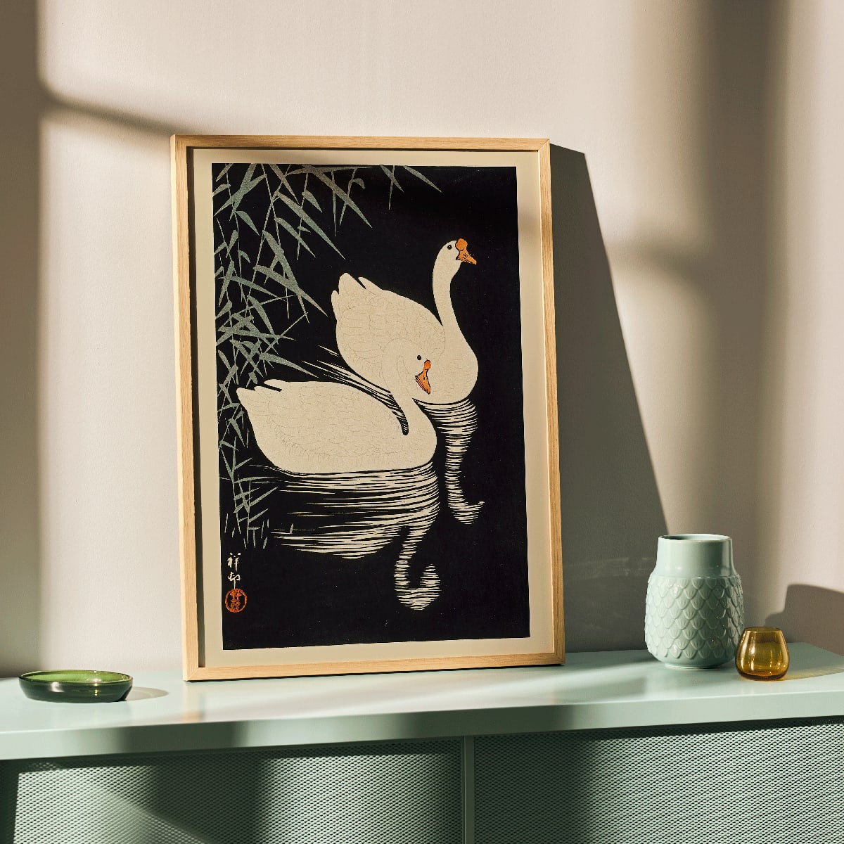 White Chinese Geese Swimming by Ohara Koson featuring two elegant white geese on water with bamboo leaves and reeds poster artwork, framed and displayed in modern living room decor